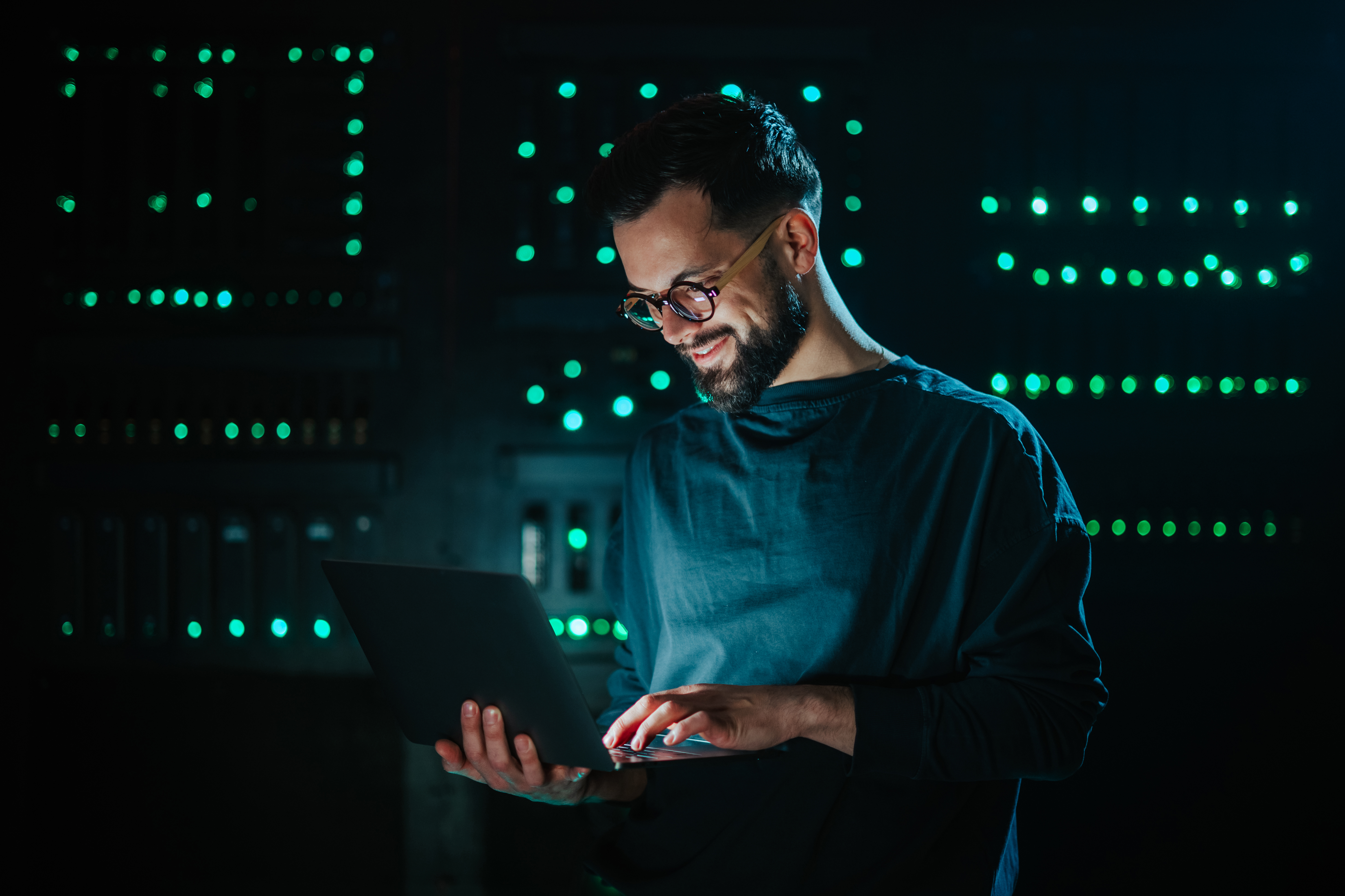 Smiling bearded man working on laptop in dark data center, managing network and database for it security